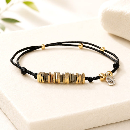 Close-up lifestyle image of a smiling blonde woman celebrating her birthday in a bright, softly lit setting. She is wearing a delicate black cord bracelet featuring stacked gold and hematite-colored milestone beads, accented with small gold spacer beads and a tiny silver charm. 