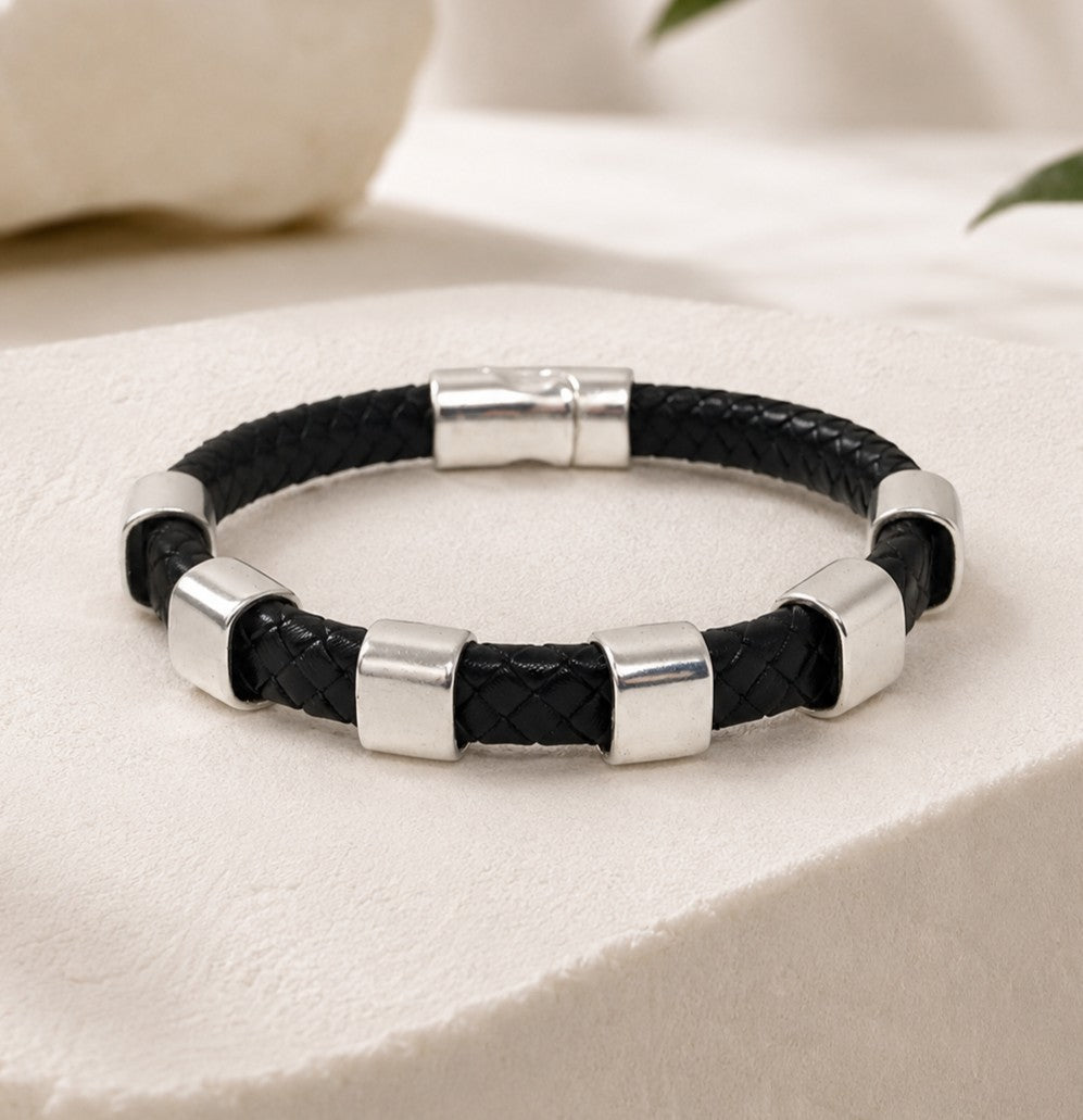 Black braided leather bracelet with polished silver-tone metal spacers and magnetic clasp. Features woven black leather cord with six rectangular metal accents evenly spaced around the bracelet. Classic masculine design with a secure magnetic closure, combining modern stainless-steel styling with textured braided leather for everyday wear. Minimalist men's bracelet suitable for casual and business-casual outfits.