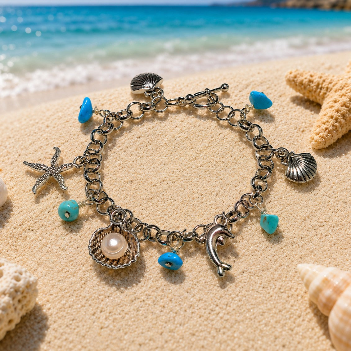 Silver-tone beach charm bracelet featuring turquoise-blue stone chip dangles, a starfish charm, seashell charms, a dolphin charm, and a pearl-in-shell charm. The bracelet has chunky linked chain construction with a toggle clasp and coastal ocean-inspired design, displayed in a circular shape.
