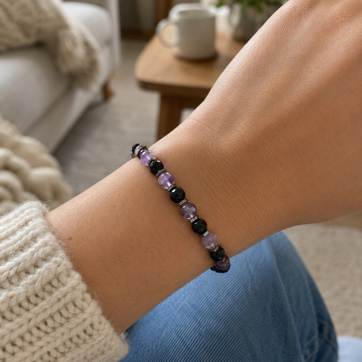 Personalized amethyst and black onyx bracelet on a woman's wrist, featuring genuine faceted purple amethyst gemstones, polished black onyx beads, and hematite spacers on an adjustable waterproof cord. This handmade crystal bracelet combines elegant purple and black gemstones for a timeless, minimalist look. Styled with a cozy cream sweater in a casual home setting, this adjustable gemstone bracelet makes a thoughtful gift for women, birthday gift, anniversary gift, bridesmaid gift, friendship jewelry.