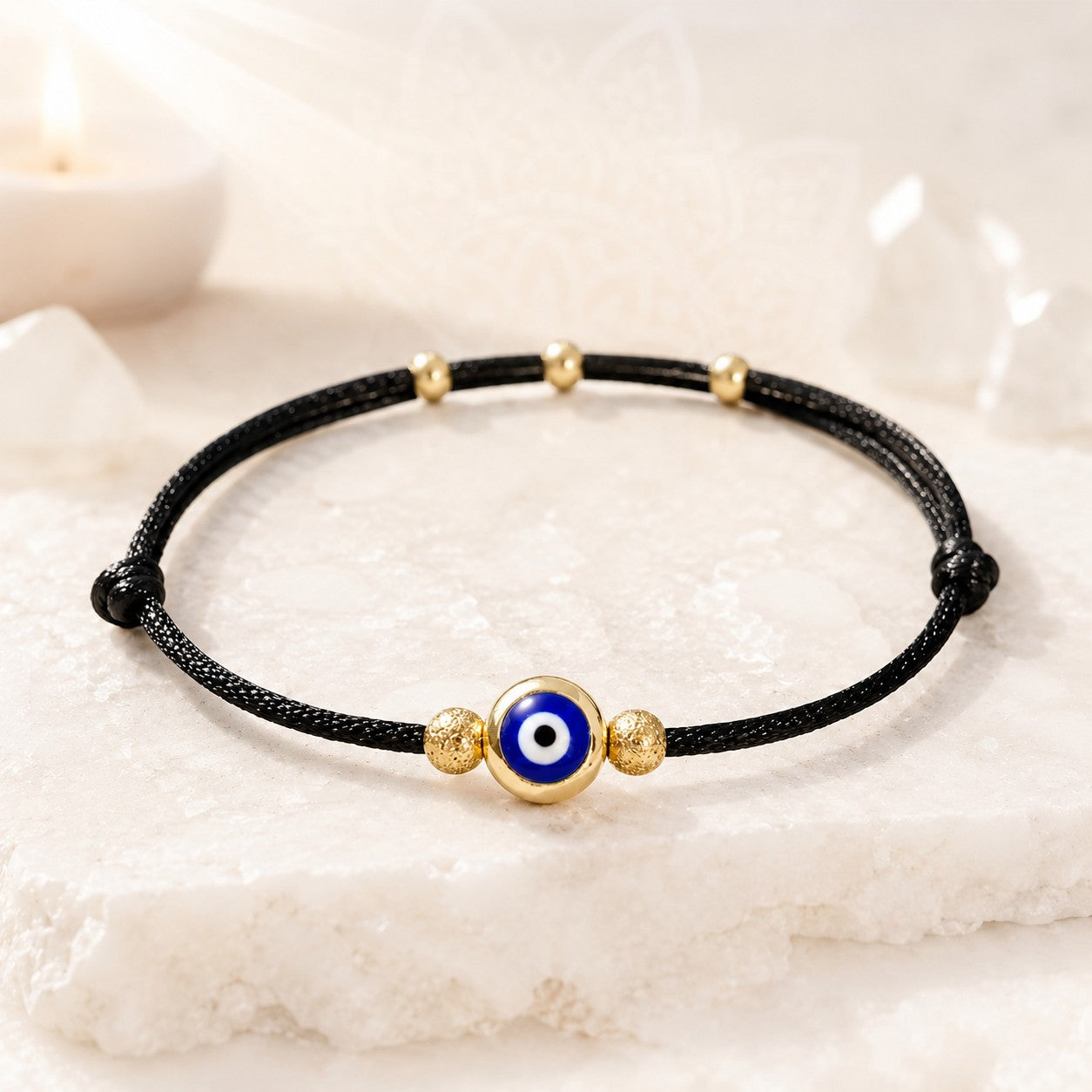 Delicate black adjustable cord bracelet featuring a small black-and-white evil eye charm framed in gold-tone metal, accented by two textured gold beads and a silver-tone initial charm. The minimalist bracelet is worn on a woman's wrist in a serene spiritual setting with soft ivory tones, natural sunlight, crystals, a candle, and subtle meditation-inspired decor in the background. The lightweight design combines meaningful symbolism with elegant everyday style, making it perfect for stacking or wearing alone