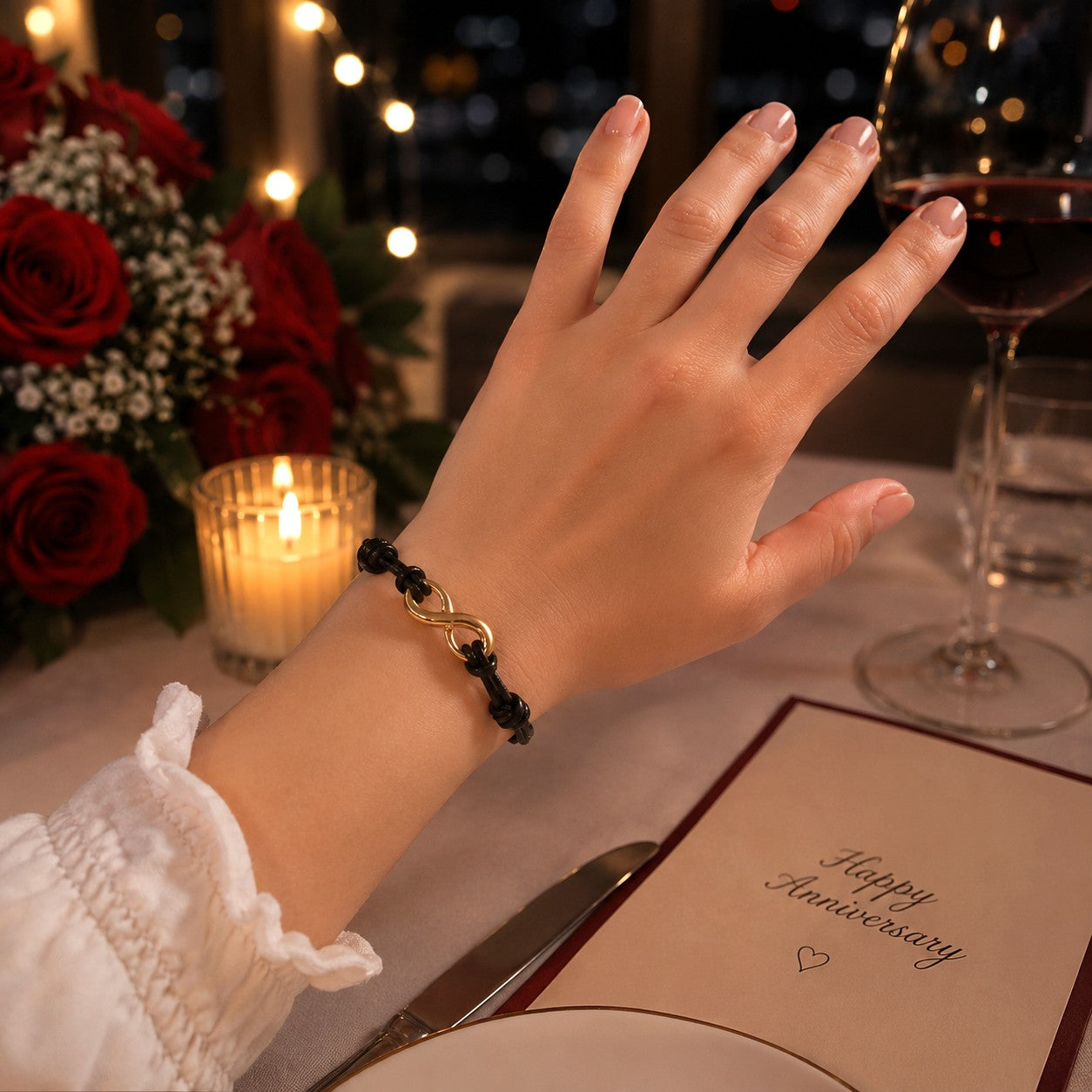 Young woman’s hand wearing a black adjustable infinity bracelet with a gold infinity charm at a romantic anniversary dinner. Elegant waterproof leather cord bracelet displayed with candlelight, red roses, wine glass, and a warm intimate restaurant setting.