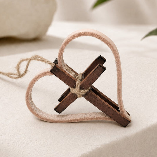 Handcrafted personalized leather and twine Christian ornament featuring a heart-shaped natural leather design intertwined with a rustic wooden cross. The cross is secured with natural jute twine and can be customized with a name, date, year, Bible verse reference, or meaningful message. Displayed on a warm neutral tabletop beside an open Bible, candle, greenery, and religious figurine, the ornament showcases its rustic farmhouse style and faith-inspired design. Made from genuine leather and twine.