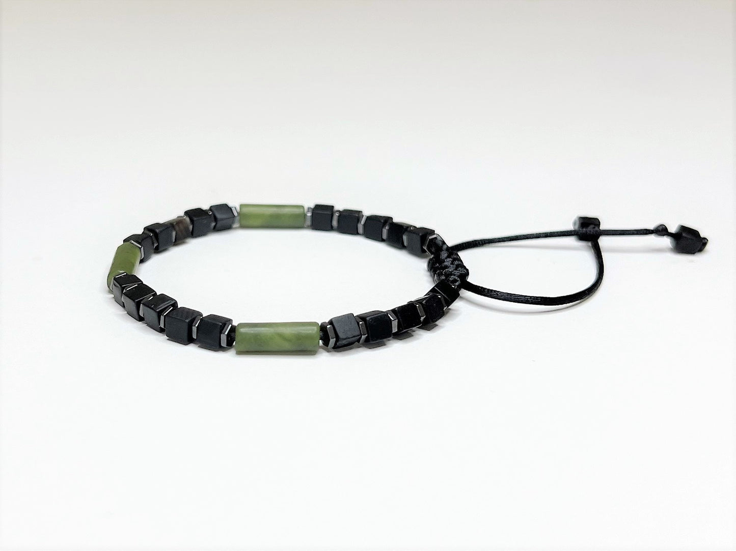 Side view of an adjustable handmade bracelet featuring a central cylindrical green jade bead flanked by matte black cubic onyx and faceted hematite spacer beads.