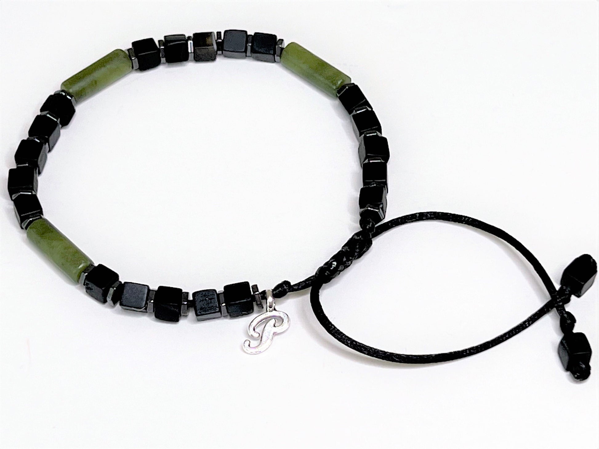 Side view of an adjustable handmade bracelet featuring a central cylindrical green jade bead flanked by matte black cubic onyx and faceted hematite spacer beads. Personalized with one initial charm.