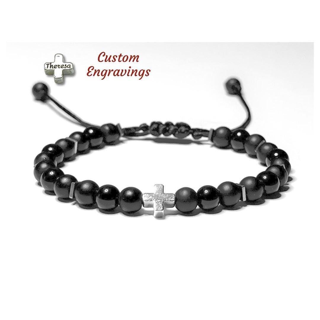 A mix of Matte and shiny black Onyx bracelet with a white gold-filled cross centerpiece.  The bracelet features round black onyx-style beads separated by small silver hematite spacers, strung on a black adjustable cord with a sliding knot closure.  The Cross can be personalized with Custom Engravings.