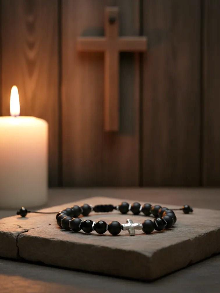 Black beaded bracelet with silver cross charm resting on a stone slab, lit by a candle in front of a wooden cross hanging on a rustic wooden wall.