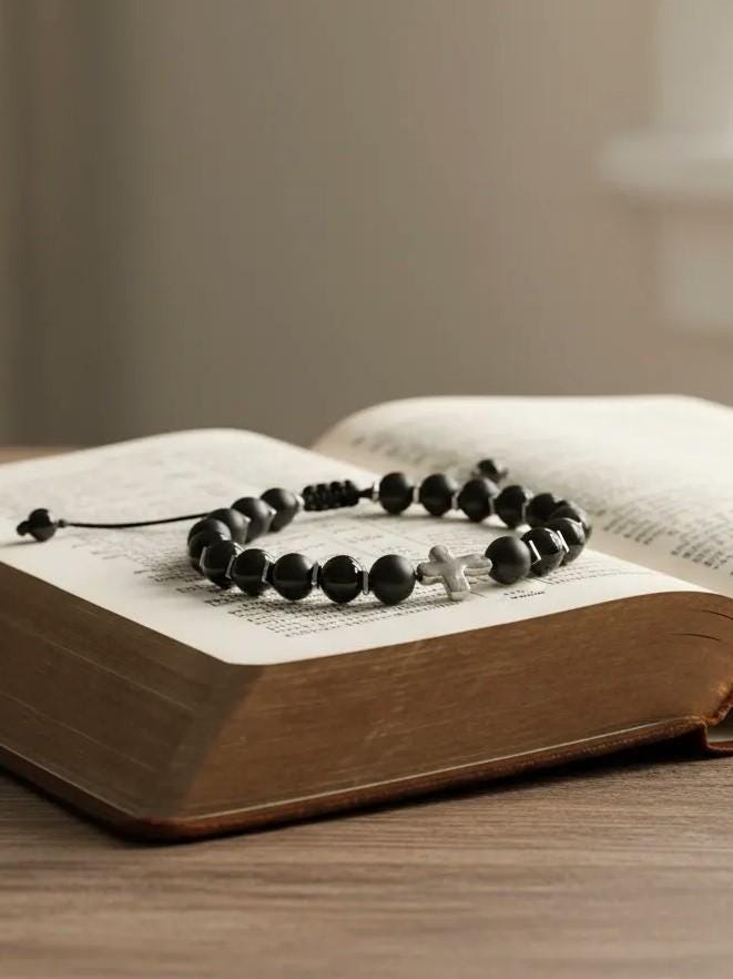 Christian bracelet made of matte black beads and a silver cross placed on the open pages of a Bible, symbolizing faith and devotion.