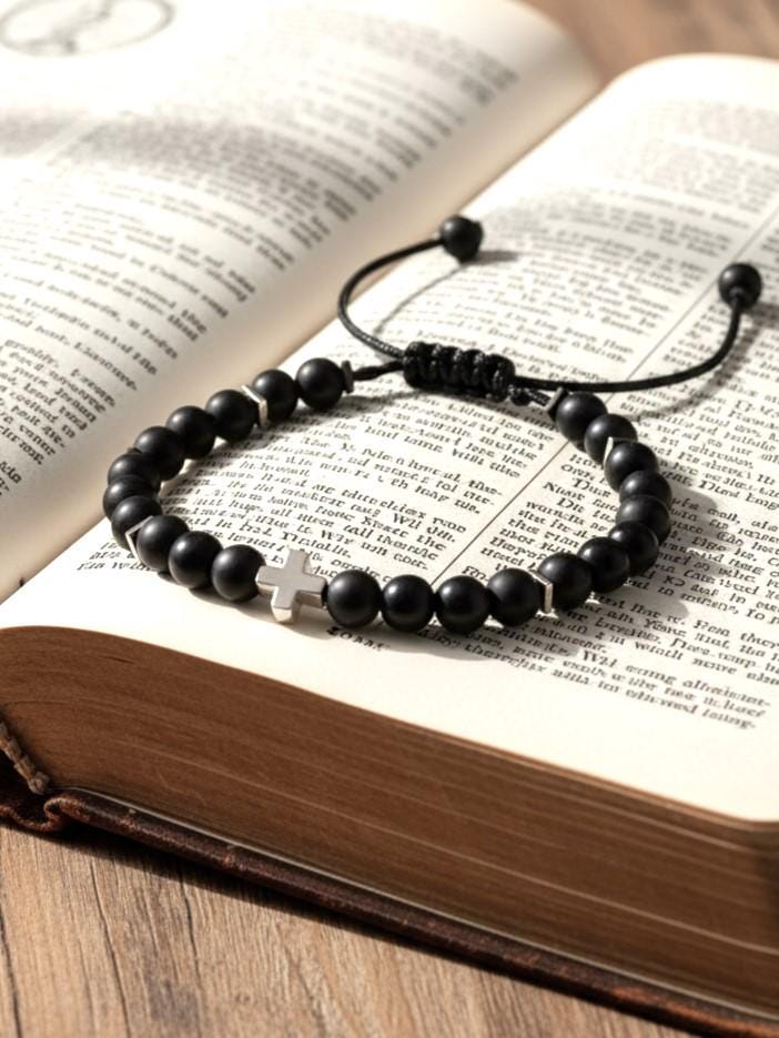 Mix of Matte and shiny black Onyx bracelet with a white gold filled cross centerpiece, resting on an open Bible. The bracelet features round black onyx-style beads separated by small silver hematite spacers, strung on a black adjustable cord with a sliding knot closure. The cross is flat and horizontally placed, visually centered on the Bible pages. The warm lighting highlights the bracelet's details and the textured wood surface beneath the book, evoking a peaceful and spiritual atmosphere.