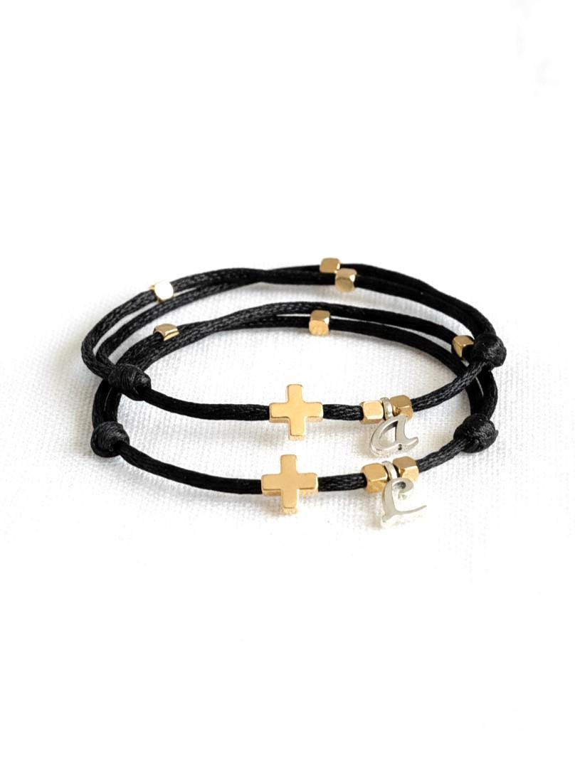 Two matching adjustable black string bracelets with 18K gold‑filled crosses and small gold accent beads, displayed on a white background. Each bracelet can be personalized with an initial charm, symbolizing faith, love, and unity. Elegant minimalist Christian jewelry for couples or families, handcrafted and waterproof for everyday wear.