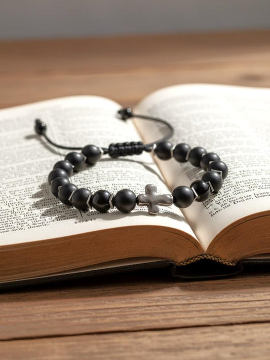 Matte black beaded bracelet with a silver cross centerpiece on a black adjustable cord, displayed on an open Bible.