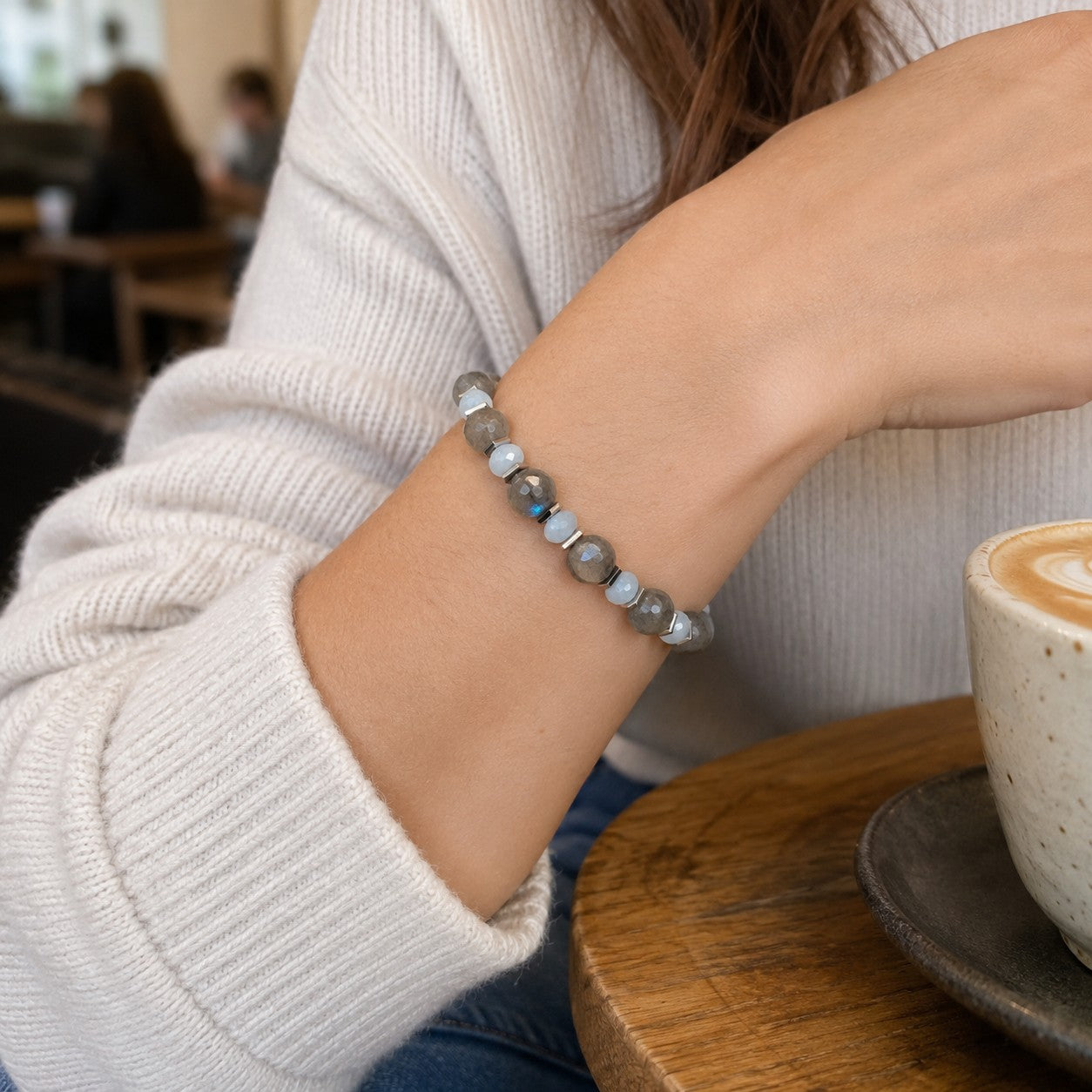 Woman wearing an adjustable Labradorite, Aquamarine, and Hematite bracelet in a cozy café setting. The handmade bracelet features larger faceted gray Labradorite gemstones, smaller round light-blue Aquamarine beads, silver-tone Hematite spacers, and a waterproof adjustable black cord. Styled with a white sweater and jeans, this elegant gemstone bracelet is perfect for everyday wear, meaningful gifts, minimalist jewelry lovers, and casual chic fashion.