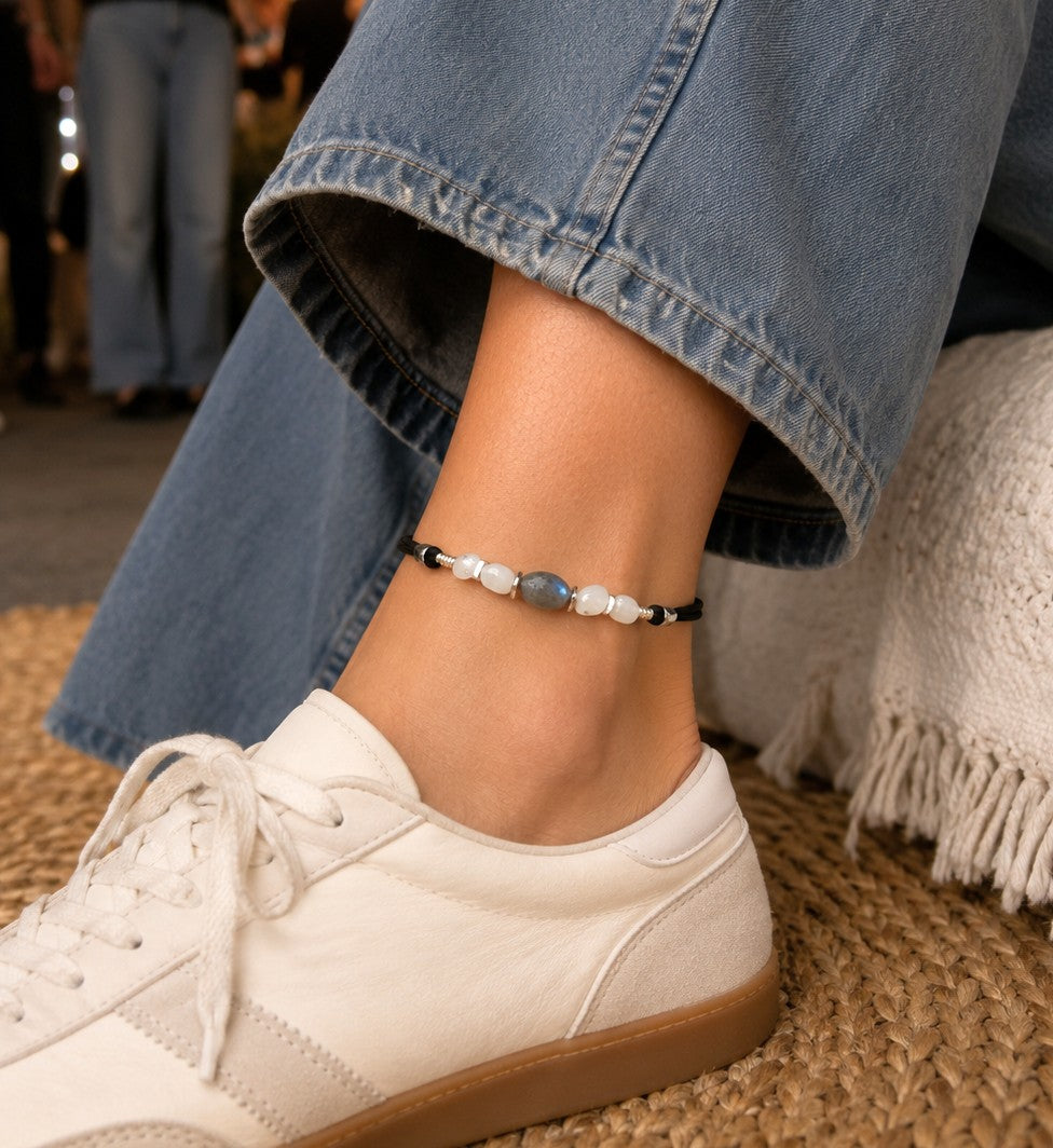 Adjustable Labradorite and Moonstone anklet on a woman's ankle at a casual party, featuring a blue-flash Labradorite gemstone, white Moonstone beads, silver-tone accents, and waterproof black cord. Styled with barrel-leg jeans and white sneakers, this handmade boho gemstone anklet is perfect for everyday wear, summer outfits, beach vacations, and unique gift ideas.