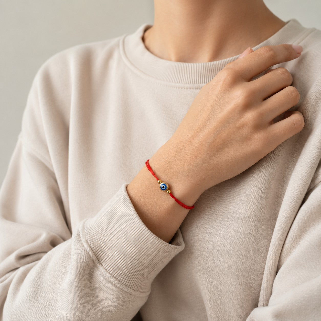 Red string evil eye bracelet on a woman’s wrist, featuring a delicate adjustable cord design with a blue protective evil eye charm and gold-tone accent beads. This minimalist handmade bracelet is worn with a casual ivory sweatshirt, creating a clean and modern everyday look. Symbolizing protection, good luck, positive energy, and spiritual well-being, this waterproof adjustable bracelet is perfect for daily wear. A meaningful gift for women, teens, daughters, sisters, best friends, mothers, or anyone who lo