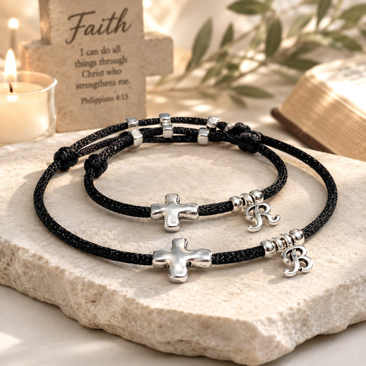 Two handcrafted silver sideways cross bracelets made with black satin cord, each featuring a silver-plated cross and a personalized initial charm—any letter can be added. One bracelet has a larger cross with a 'B' charm, the other has a smaller cross with an 'R' charm. Adjustable, waterproof, and gift-ready, displayed on a white background.