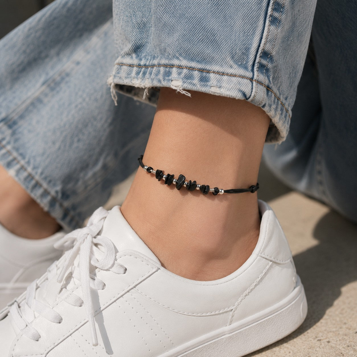 Close-up lifestyle photo of a black tourmaline Gothic anklet worn around a woman’s ankle. The handmade anklet features polished black tourmaline chip gemstones accented with silver-tone spacer beads and strung on an adjustable black cord. The model is dressed in relaxed wide-leg light-wash denim jeans paired with clean white tennis shoes, creating a casual everyday look. 