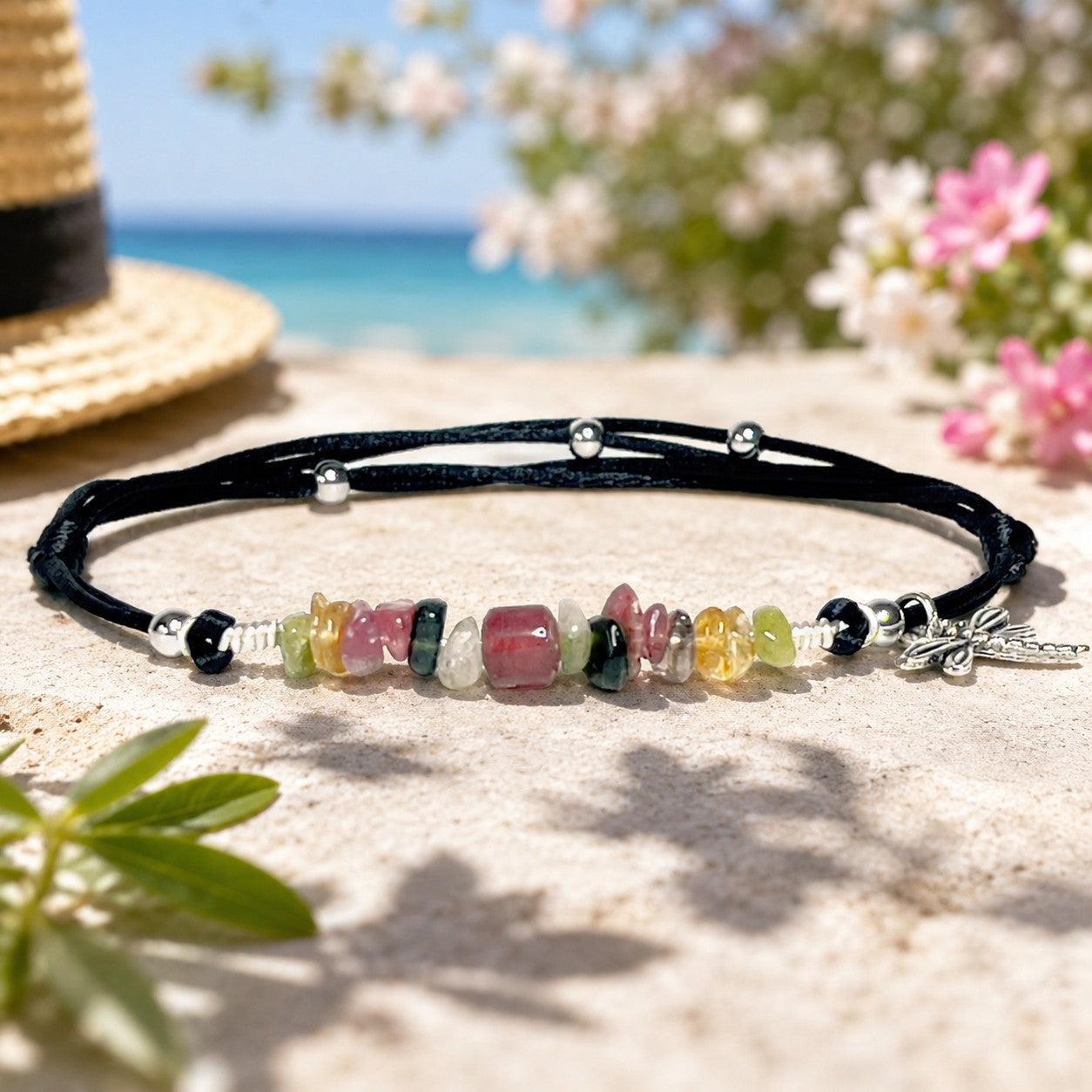 A handmade anklet with small round multicolor tourmaline beads in shades of pink, green, and black, accented by tiny silver-grey hematite squares. The beads are strung on a thin black nylon cord with an adjustable sliding knot. A delicate silver dragonfly charm hangs near the clasp, adding a playful, lightweight touch. The anklet has a casual, boho style and comes presented in a simple gift box.