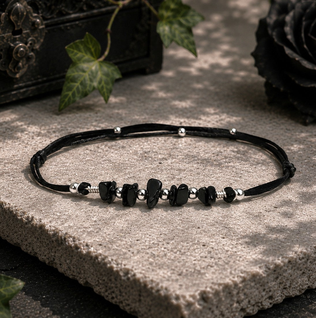 Gothic Black tourmaline gemstone bracelet displayed on a textured stone slab in a softly lit gothic-inspired setting. The adjustable bracelet features a black cord with natural black tourmaline chip stones accented by polished silver-toned beads and decorative metal spacers. The bracelet is arranged in an oval shape and centered on a light gray stone platform. The image emphasizes the bracelet's handcrafted design, natural stone textures, and minimalist aesthetic against a moody yet refined backdrop.