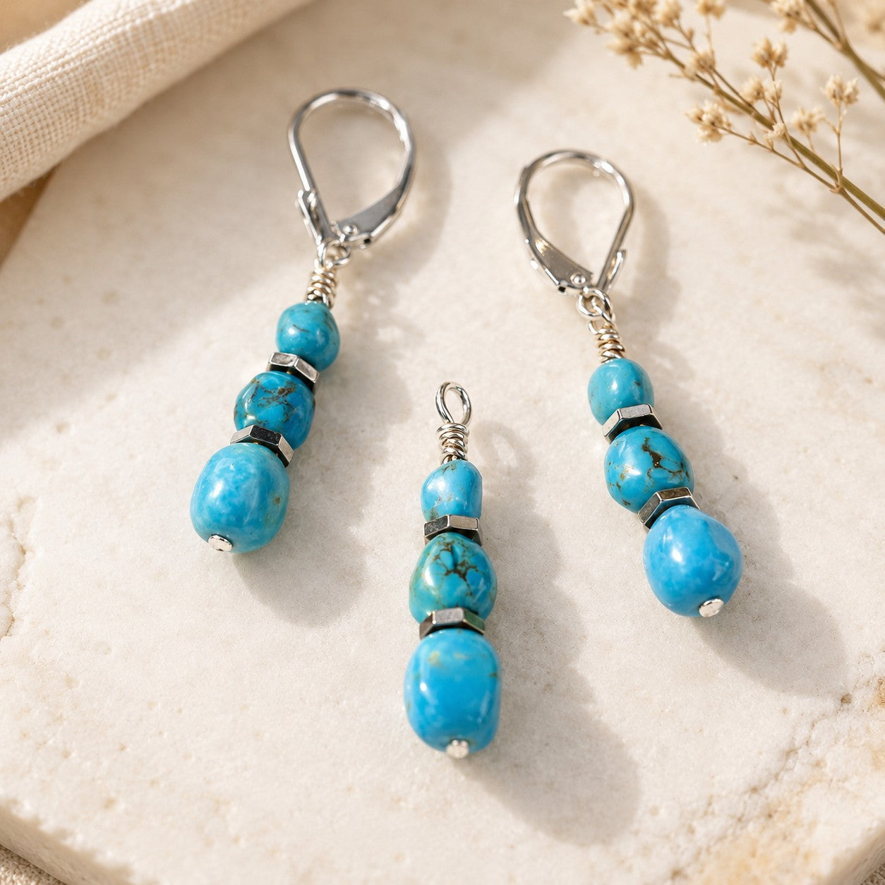 Detailed product photo of a handmade turquoise jewelry set featuring matching dangle earrings and a coordinating pendant. The set includes vibrant blue turquoise gemstone beads with natural dark matrix veining, accented by polished silver-tone spacer beads and wrapped wire details. Two leverback earrings frame the center pendant, which is displayed without a chain to highlight the beadwork design. 